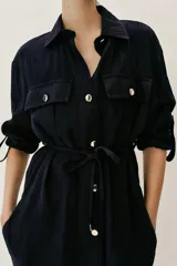 Camisa estilo safari de color azul marino con textura efecto arrugado, cuello solapa, mangas remangadas y cierre frontal con botones. Presenta bolsillos delanteros con solapa y un cinturón fino ajustable en la cintura.