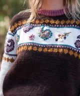 Sweater de punto en lana merino, con diseño de bloques de color en marrón, crema y fucsia. Presenta detalles de bordados artesanales y motivos jacquard en el pecho y las mangas, cuello redondo y mangas largas.