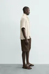 Camisa de calce relajado confeccionada en tejido con algodón de color beige. Presenta cuello bowling, manga corta, aberturas laterales en el bajo y cierre frontal con botones.