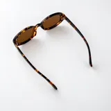 Lentes de sol con armazón ovalado de carey y micas marrones, con detalle metálico dorado en las esquinas y patillas.