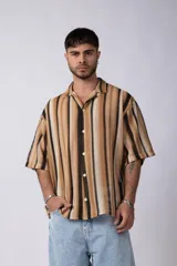 Camisa de manga corta con cuello solapa y cierre frontal de botones. Presenta un diseño de rayas verticales en tonos marrones, beige y negro, con una textura tejida y corte holgado.