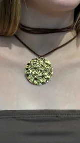 Gargantilla tipo choker con cordón de gamuza marrón y dije circular grande de metal dorado con textura irregular y efecto martillado.