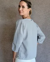 Blusa de crepe color gris claro, con mangas tres cuartos que presentan cortes y pespuntes decorativos. Posee un diseño holgado y cuello redondo.