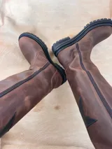 Botas de cuero color marrón con caña alta, suela track y cierre lateral con cremallera. Detalle de estrella en cuero negro en la parte superior de la caña.