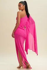 Vestido largo de fiesta color fucsia, con diseño de un solo hombro y manga tipo capa fluida. Presenta un detalle de fruncido lateral con aplique metálico en la cintura y falda con tajo lateral.