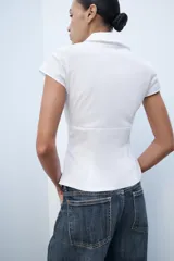 Top de popelina blanca de corte corsetero, con escote en pico pronunciado, cuello tipo solapa, mangas muy cortas y costuras marcadas.