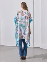 Kimono ligero de manga corta, color blanco, con estampado floral estilo cachemira en tonos pastel (naranja, verde menta, celeste). Presenta bordes y detalles en azul turquesa con borlas colgantes en el ruedo.