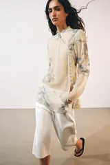 Camisa de cuello redondo y manga larga con diseño de estampado artístico en tonos azules sobre fondo beige. Presenta un cierre frontal asimétrico con botones forrados a tono y una abertura lateral en el escote.