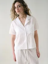 Camisa de manga corta para dormir confeccionada en broderie de algodón, con cuello solapa y cierre frontal con botones. Presenta un bolsillo aplicado en el pecho.