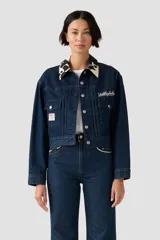 Chaqueta vaquera azul oscuro con cuello estampado de vaca en blanco y negro. Presenta bordado blanco en el pecho izquierdo con la palabra "Yodelayhehoo" y un parche con el logo de Levi's en el bolsillo derecho.