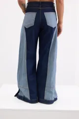 Pantalón wide leg de tiro alto confeccionado en denim de diseño bicolor, combinando tonos azul oscuro y celeste con costuras en contraste. Presenta bolsillos tipo parche en la parte inferior de las piernas y aberturas en los puños.