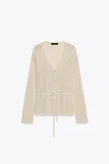 Chaqueta de punto color beige claro, con escote en V, manga larga y cierre frontal de botones. Presenta un cinturón fino del mismo tejido anudado a la cintura.
