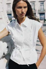 Camisa blanca de popelín con cuello solapa, manga sisa y detalle de guipur floral en el frente. Incluye un cinturón fino del mismo tejido que se ata en la cintura.