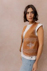 Chaleco corto de efecto gamuza color tostado, con detalles bordados en contraste en los bordes y en la parte inferior. Presenta escote en V y diseño abierto al frente.
