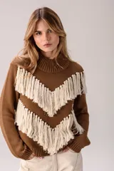 Sweater de punto color marrón con cuello medio alto y silueta relajada. Presenta un diseño distintivo con líneas en V decoradas con flecos largos de color crema aplicados a mano.