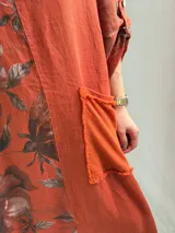 Vestido largo color terracota con diseño asimétrico que incluye un lateral con estampado floral grande. Presenta mangas tres cuartos, escote redondo y un bolsillo aplicado en el frente.
