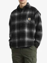 Camisa tipo sobrecamisa Carhartt de corte holgado, confeccionada en tejido de lana a cuadros en tonos negro y gris. Presenta cierre frontal de botones a presión, cuello clásico, mangas largas con puños abotonados y un bolsillo en el pecho con etiqueta de la marca.