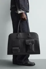 Bolso de viaje negro con acabado grabado y diseño de múltiples bolsillos frontales. Dispone de un compartimento principal con cierre magnético, un bolsillo interno con cremallera y tres bolsillos exteriores de diversos tamaños con cierre de imán. Incluye dos asas de mano superiores.
