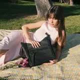 Cartera bandolera de símil cuero negro, de formato amplio y flexible. Cuenta con cierre superior, bolsillo frontal con cierre y detalle decorativo colgante, bolsillo trasero y correa larga ajustable y desmontable.