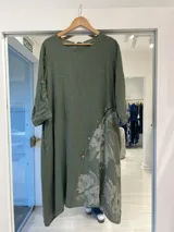 Vestido midi de lino color verde militar, con diseño holgado, cuello redondo y mangas largas con presilla para arremangar. Cuenta con un corte diagonal cruzado decorado con botones y un estampado floral difuminado en la parte inferior.