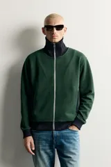 Chaqueta regular fit color verde oscuro con detalles en negro en el cuello subido, los puños, el dobladillo inferior y los ribetes de los bolsillos. Cuenta con cierre frontal de cremallera metálica y bolsillos con cierre en la cadera.