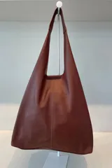 Bolso tipo tote de gran capacidad, confeccionado artesanalmente en cuero vacuno color marrón. Presenta un diseño minimalista con costura central vertical, fuelle lateral ajustable con cierre y placa metálica con logo en la parte inferior.