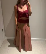 Pollera larga color marrón claro con textura, corte amplio y talle alto. Presenta detalles de ribete brillante color rojo/bordó en los paneles frontales.