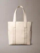 Cartera tote Calvin Klein de color beige, confeccionada en suave piel sintética, con diseño estructurado, asas fijas y compartimentos exteriores en los laterales. Presenta detalles de cinta con logo en jacquard y logo metálico grabado en la parte delantera.