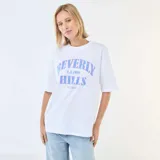 Camiseta de corte oversize color blanco, con cuello redondo y mangas cortas. Presenta un estampado frontal con la inscripción 'Beverly Hills L.A. 1989 West Co' en tonos azules.
