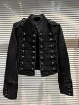 Chaqueta corta de denim negro con diseño de inspiración militar, cuello mao, cierre frontal con múltiples botones metálicos y detalles de trenzado horizontal. Presenta charreteras en los hombros y puños con botones decorativos.