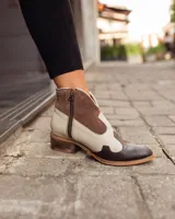 Botinetas estilo western de caña baja, confeccionadas en una combinación de cuero liso y gamuza en tonos marrón oscuro, marrón medio y crema. Presentan cierre lateral, taco bajo cuadrado y diseño con recortes característicos del estilo cowboy.