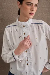 Camisa de popelina blanca con estampado de motivos deportivos, cuello alto con volados, cierre frontal con botones y mangas largas con puños elásticos y volados.