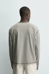 Camiseta de manga larga con cuello panadero (henley) y cierre de tres botones. Presenta un patrón de rayas horizontales finas en tonos beige y negro, con pespuntes a contraste en el cuello, puños y bajo.