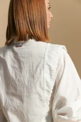 Camisa blanca de manga larga con cuello y botones frontales. Presenta detalles de volados o puntilla en los hombros y mangas abullonadas con puños anchos. La tela parece tener un sutil tejido a rayas verticales.