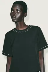 Top negro de manga corta con cuello redondo, adornado con perlas blancas aplicadas en el escote y en los puños de las mangas. Cierre posterior con botones de perla.