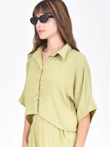 Camisa de corte cropped y holgado, color verde oliva, con cuello clásico, cierre frontal de botones y mangas cortas con dobladillo.