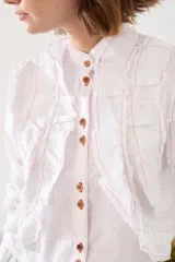 Camisa de lino blanca con cuello mao, manga larga y botones frontales. Presenta volados delanteros con terminaciones desflecadas y detalles de costuras en hilo rojo.