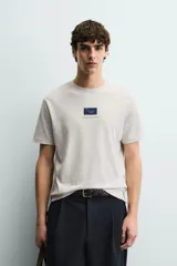 Camiseta de corte relajado, color gris jaspeado, con cuello redondo y manga corta. Presenta un parche rectangular azul marino en el centro del pecho con texto estampado en blanco y una frase adicional debajo.