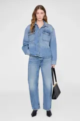 Camisa de denim en tono azul medio con corte boxy y estilo workwear. Presenta cuello clásico, cierre frontal con botones a presión, hombros caídos y dos bolsillos de parche en el pecho.