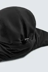 Gorra técnica de color negro con diseño de cinco paneles y orejeras integradas ajustables mediante una trabilla superior. Confeccionada en tejido acolchado, cuenta con ajuste elástico en la parte posterior.