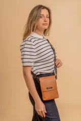 Phonebag pequeña color mostaza, confeccionada en cuero sintético, con solapa frontal y correa ajustable con logo de la marca.