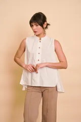 Blusa sin mangas de color blanco, con cuello mao y cierre frontal de botones. Presenta un diseño holgado y asimétrico, con la parte trasera más larga y aberturas laterales en el dobladillo.