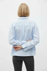 Camisa de algodón a rayas celestes y blancas, de corte holgado, con cuello camisero, cierre frontal de botones y mangas largas con puños rectos.