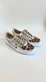 Sneakers de mujer en tonos blancos, dorados y detalles con estampado animal print de leopardo en la puntera. Incorporan cordones frontales y apliques metálicos decorativos.