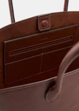 Bolso tipo tote marrón de poliuretano, de gran tamaño, con dos asas y cierre de botón magnético. Detalle de costuras visibles y laterales ajustables.