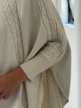 Poncho de tejido de punto color beige con cuello alto y diseño de trenzas verticales en el frente. Presenta mangas integradas con puños acanalados y un corte holgado con ruedo redondeado.