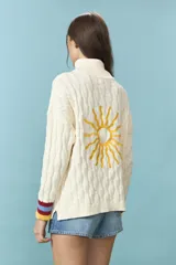 Sweater de tejido de punto con estructura de ochos en color natural, de cuello alto y manga larga. Presenta un bordado de sol en la espalda y puños con franjas de colores en rojo, celeste y amarillo.
