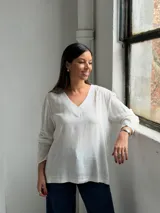 Blusa blanca de corte holgado con escote en V, mangas largas y costuras visibles en el cuello y el ruedo.