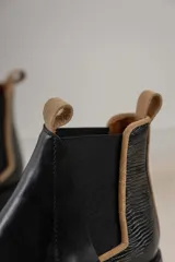 Botas tipo Chelsea de cuero negro, con paneles laterales elásticos y detalle de textura grabada en el talón. Presentan un borde en color beige y tiradores en la parte superior.
