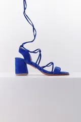 Sandalia de taco cuadrado medio, confeccionada en cuero gamuzado color azul vibrante. Presenta un diseño de tiras finas que se entrelazan y se atan alrededor del tobillo, con puntera abierta.